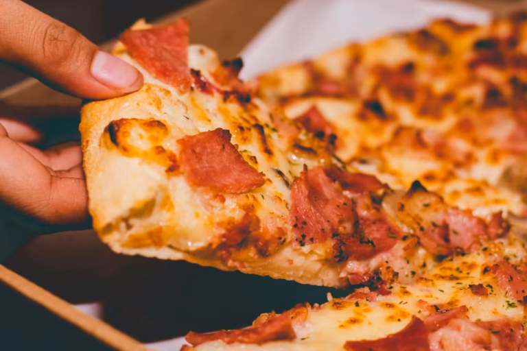 Kawałek pizzy z szynką i serem trzymany w dłoni. Widoczne grube, puszyste ciasto, stopiony ser, plastry szynki i zielone zioła. Zdjęcie do artykułu analizującego potencjalne zagrożenia i korzyści zdrowotne związane ze spożyciem pizzy.